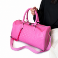 A bright pink crocodile-embossed duffel bag with dual top handles and a matching shoulder strap, held by a person wearing a black top and white pants against a white background.