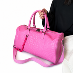 A bright pink crocodile-embossed duffel bag with dual top handles and a matching shoulder strap, held by a person wearing a black top and white pants against a white background.