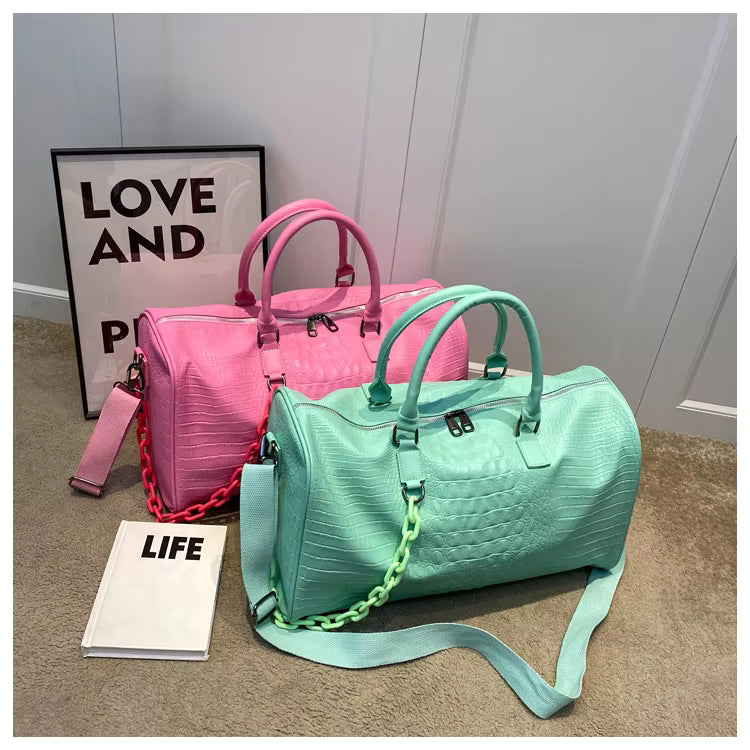 Two embossed faux-croc duffle bags displayed on a carpeted floor: one in bright pink and one in mint green, each with matching shoulder straps and chain details, styled with a framed 'LOVE AND' sign and a book labeled 'LIFE'.