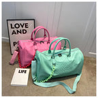 Two embossed faux-croc duffle bags displayed on a carpeted floor: one in bright pink and one in mint green, each with matching shoulder straps and chain details, styled with a framed 'LOVE AND' sign and a book labeled 'LIFE'.