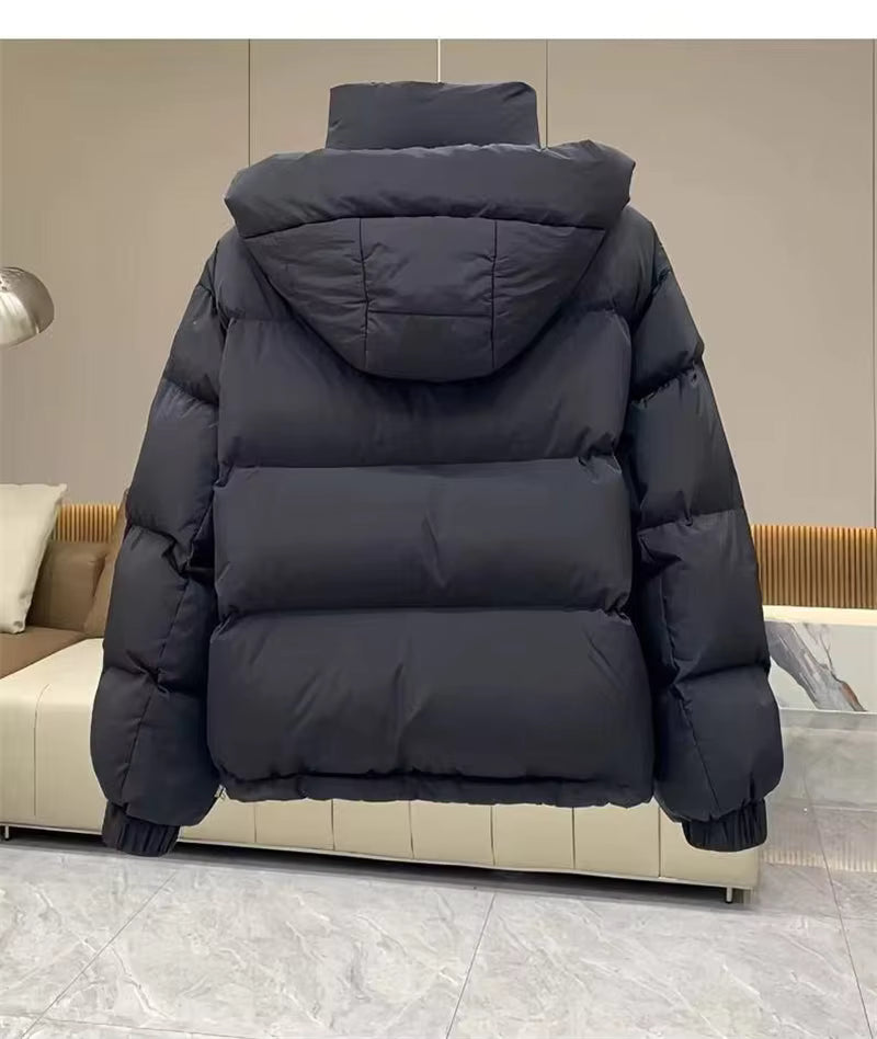 A black hooded puffer jacket shown from the back, resting on a modern beige sofa in an indoor setting with gray tiled flooring.