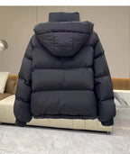 A black hooded puffer jacket shown from the back, resting on a modern beige sofa in an indoor setting with gray tiled flooring.