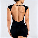 Rear view of a woman wearing a black open-back bodysuit with cap sleeves and a small gold clasp at the neckline, against a light gray studio background.