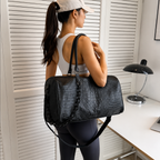 A woman in athletic wear carries a large black crocodile-embossed duffel bag with a shoulder strap in a bright, modern room with white louvered doors and a desk.